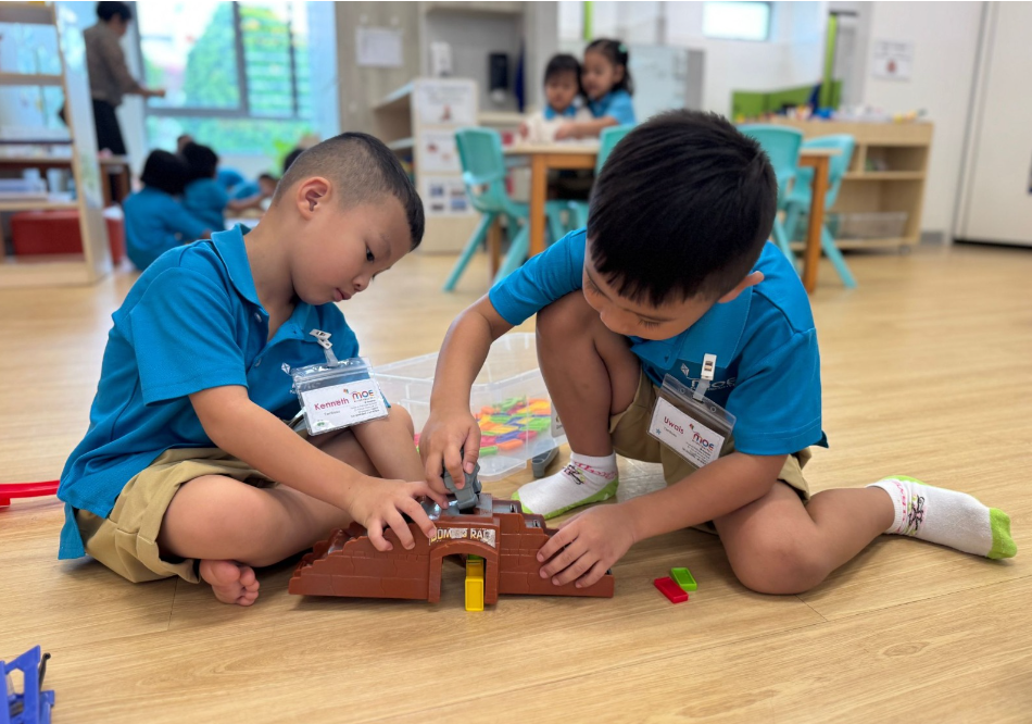 Two children in blue shirts sit on a classroom floor, playing with a brown toy ramp and blocks.